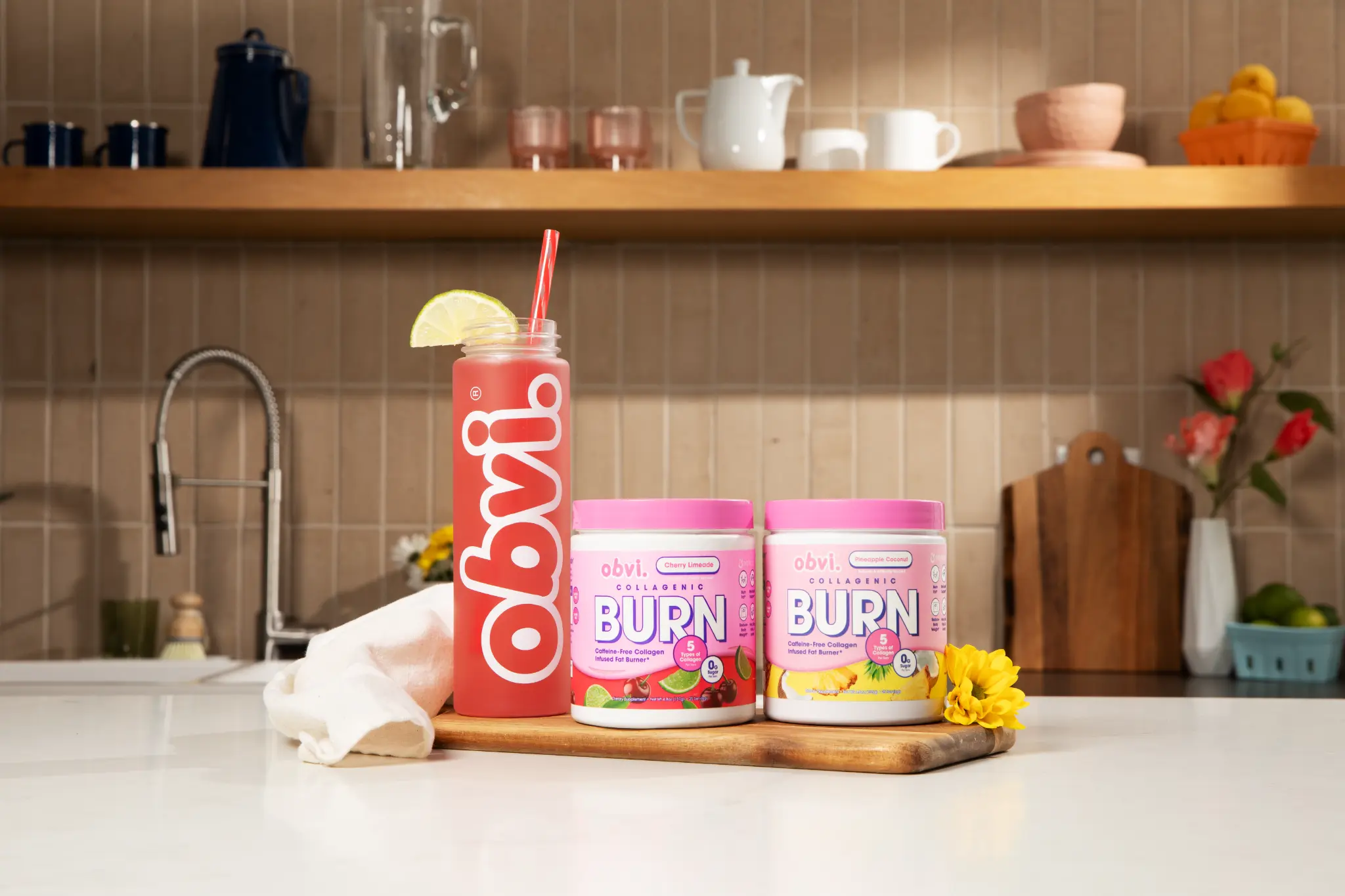 Obvi Collagenic Burn Powder two tubs and bottle on kitchen counter with yellow flowers